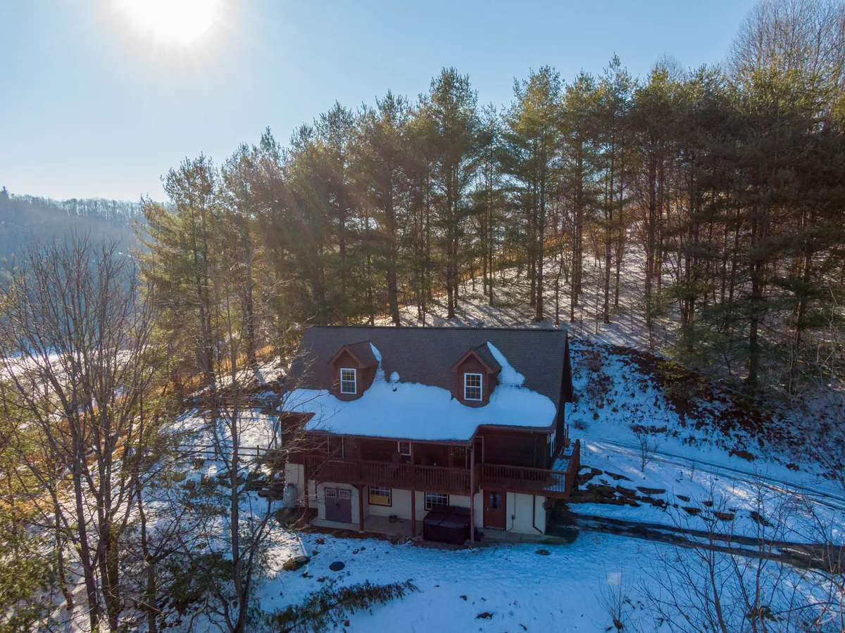 A wide exterior shot of the house in winter, with a snow-covered roof and trees in the background. The scene evokes the unique winter charm of the setting and hints at the adventure of accessing the property via snow-covered roads, subtly referencing the need for 4WD/AWD vehicles. The porch and surrounding area invite cozy winter activities, such as sipping cocoa or watching the sunrise.