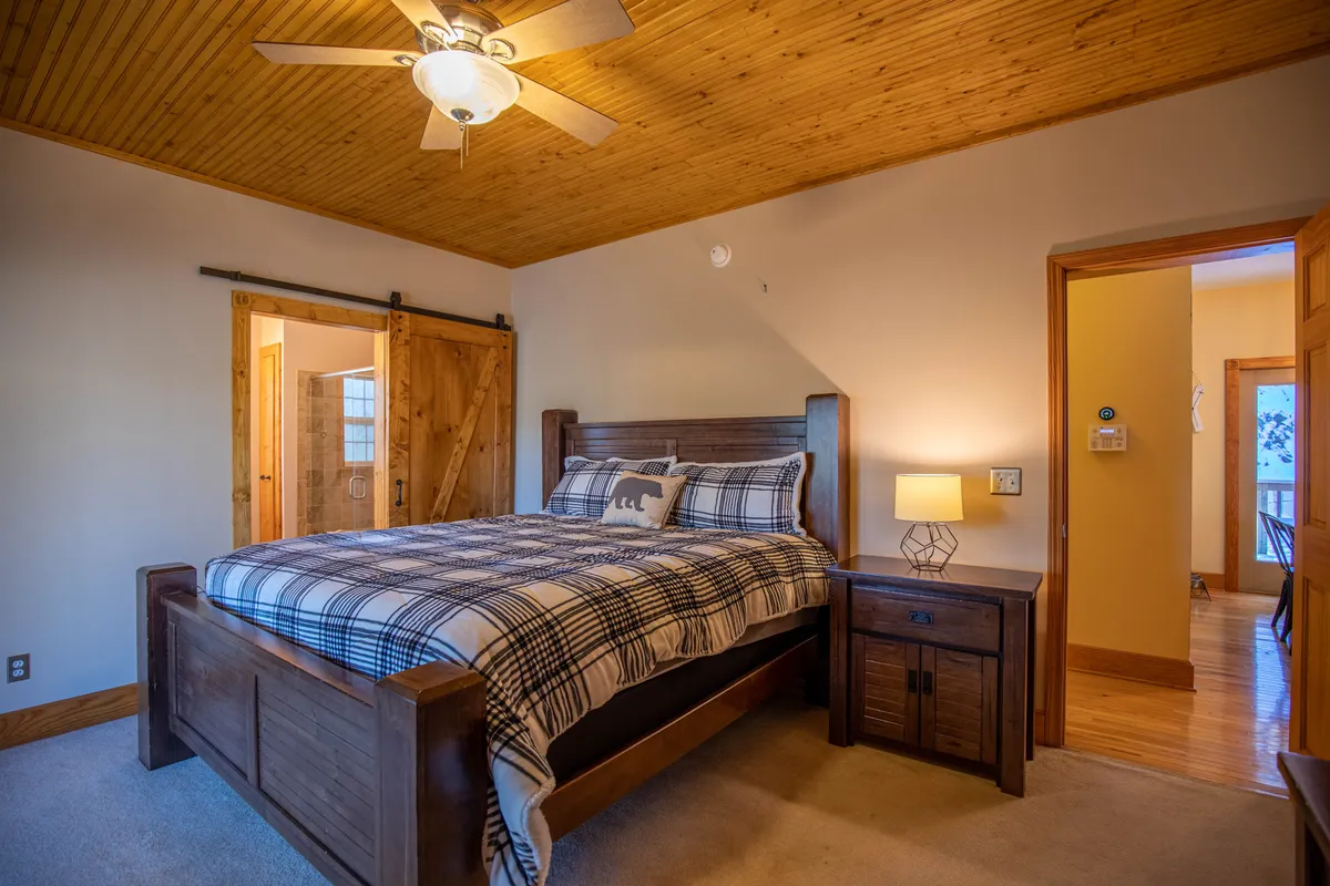 This image depicts a tranquil bedroom featuring a wooden bed frame with cozy plaid bedding, a nightstand with a lamp, and a barn-style door. Sunlight streams through the window, creating a calming and restful atmosphere that invites relaxation.