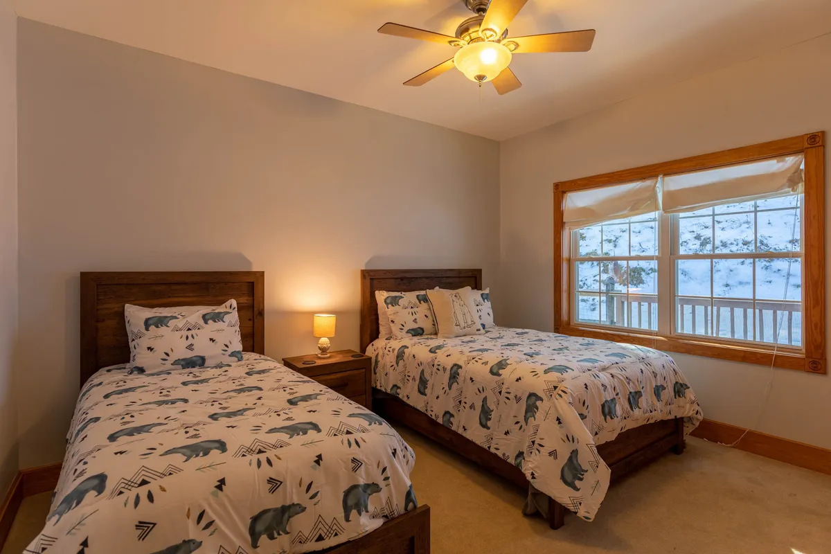 This bright bedroom features two beds with patterned bedding, a window offering a soothing view of nature, and a nightstand with a lamp. The space is airy and inviting, providing ample room for privacy and relaxation for guests or family.