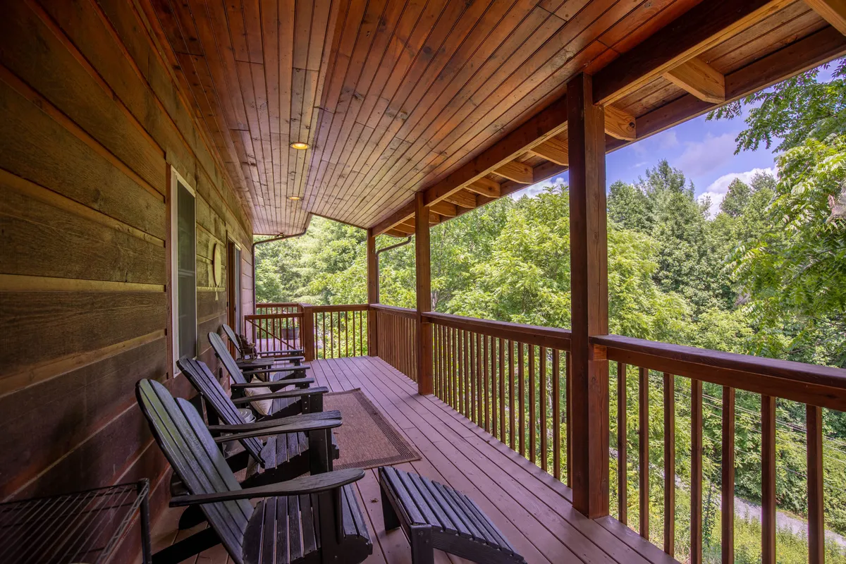 The wooden deck is adorned with classic rocking chairs, set against a lush backdrop of greenery. The space is ideal for relaxing, socializing, or quietly enjoying the surrounding natural landscape. The tranquil mood is enhanced by the gentle play of light and the inviting arrangement of furniture.