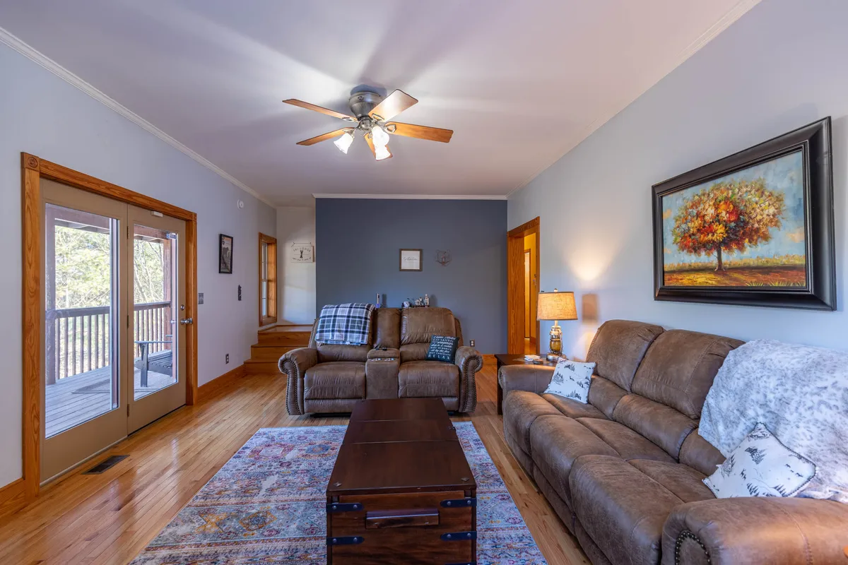 A family-oriented living space with comfortable seating arranged around a wooden coffee table, decorative painting on the wall, and large windows leading to a porch. The layout invites both quiet moments and lively gatherings.