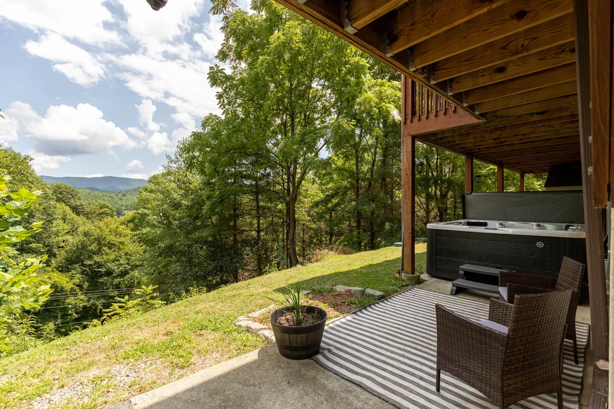 A striking exterior shot of a wooden deck featuring a hot tub and comfortable seating area, all oriented toward expansive mountain views. The setting glows with soft evening light, suggesting opportunities for both daytime relaxation and nighttime stargazing. The surrounding greenery and peaks frame the space in natural beauty.